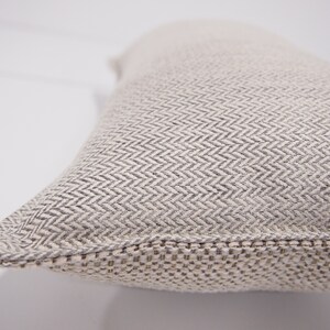 May include: A close-up of a beige pillow with a subtle herringbone pattern. The pillow has a woven edge.