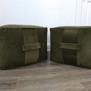 May include: Two green velvet square ottomans with a single stitched seam down the center of each side. Each ottoman has a fabric handle on the side.