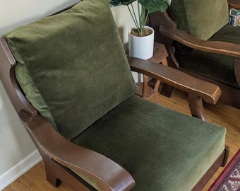 Könnte beinhalten: Zwei Vintage-Sessel mit dunklen Holzrahmen und olivgrünen Samtkissen. Die Stühle haben ein klassisches Design mit geschwungenen Armlehnen und einer hohen Rückenlehne. Zwischen den Stühlen steht ein kleiner Beistelltisch mit einer Topfpflanze.