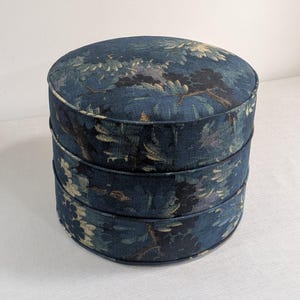 May include: A round, stacked ottoman featuring a dark blue fabric upholstery with a repeating floral and foliage pattern in shades of light blue, cream, and black. The cylindrical piece has visible piping details separating the three stacked sections.