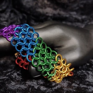 Pode incluir: Um bracelete de malha de corrente arco-íris feito de anéis de metal. O bracelete é feito nas cores do arco-íris, começando com roxo e terminando com dourado.