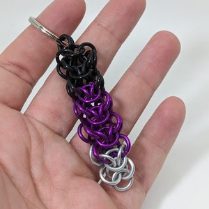 Può includere: Un portachiavi in maglia di catena nero, viola e argento. Il portachiavi è realizzato con piccoli anelli di metallo collegati tra loro in un motivo a maglia di catena. La parte superiore del portachiavi ha un anello argentato per fissarlo a un portachiavi.