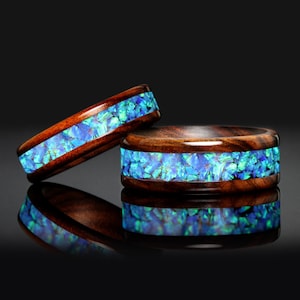 May include: Two wooden rings featuring a blue opal inlay. The rings are crafted from dark brown wood with a polished finish. The opal's shades of blue and green provide a striking contrast against the wood's grain.