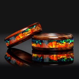 May include: Two wooden rings with a vibrant inlay of colourful opal fragments. The rings feature a rich, dark wood exterior and a dazzling display of orange, yellow, green, and blue within the band. The rings are displayed on a reflective black surface.