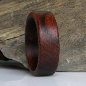 Puede incluir: Un anillo de madera hecho a mano, de color marr&oacute;n oscuro con vetas visibles. El anillo es liso y pulido, mostrando la belleza natural de la madera. El anillo tiene un dise&ntilde;o de banda simple, ideal para el uso diario.