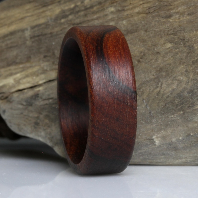Puede incluir: Un anillo de madera hecho a mano, de color marr&oacute;n oscuro con vetas visibles. El anillo es liso y pulido, mostrando la belleza natural de la madera. El anillo tiene un dise&ntilde;o de banda simple, ideal para el uso diario.