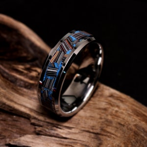 May include: A silver ring with a mosaic inlay of blue, black, red, and white stripes. The ring is displayed on a wooden surface. The ring's design is eye-catching and modern.