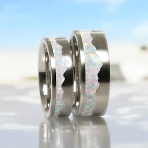 May include: A pair of silver rings with a mountain range design. Each ring has a white, textured inlay with iridescent specks. The rings are set against a blurred blue and white background.