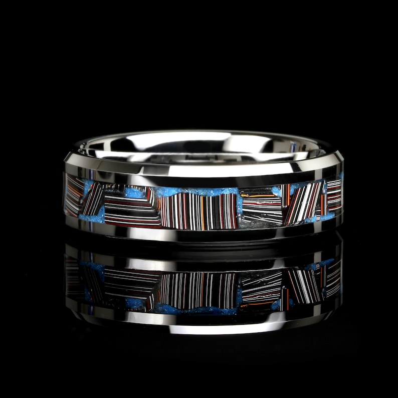 May include: A silver ring with a distinctive design. The ring has a band of colourful, rectangular inlays set against a light blue background. The inlays feature a striped pattern with red, black, and white lines. The ring is displayed on a black surface.