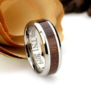 May include: A silver ring with a dark brown wood inlay. The ring has the engraved text "Team TNI 2019" on the inside. The ring is set against a blurred background of a flower.