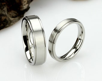 Könnte beinhalten: Zwei silberne Eheringe mit gebürsteter Oberfläche. Die Ringe haben einen leicht erhöhten Rand.