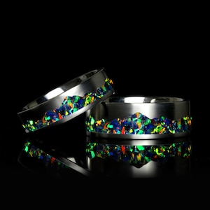 May include: Two silver rings with a band of iridescent green, blue, and red opal stones. The rings are stacked on top of each other.