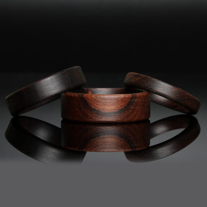 Puede incluir: Cuatro anillos de madera de diferentes anchos se muestran sobre un fondo oscuro. Los anillos est&aacute;n hechos de madera marr&oacute;n oscuro con patrones de vetas visibles. Los anillos est&aacute;n dispuestos para mostrar su dise&ntilde;o y textura.