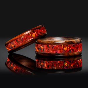 Könnte beinhalten: Zwei Holzringe mit einem lebendigen roten und orangen Inlay. Die Ringe haben eine dunkle Holzaußenseite und eine farbenfrohe, strukturierte Innenseite. Die Ringe werden auf einer reflektierenden schwarzen Oberfläche präsentiert, die ihr aufwendiges Design und ihre Handwerkskunst zeigt.