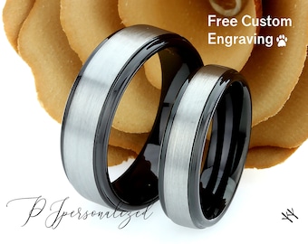 May include: Two wedding rings with a brushed silver center and black edges. The rings are set against a blurred background of a tan flower. The text "Free Custom Engraving" is visible.