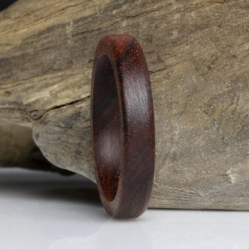 Puede incluir: Un anillo de madera marr&oacute;n oscuro con un acabado liso y pulido. El anillo est&aacute; hecho de una rica madera marr&oacute;n rojiza y tiene un dise&ntilde;o simple y cl&aacute;sico. El anillo mide aproximadamente 2 cm de ancho.