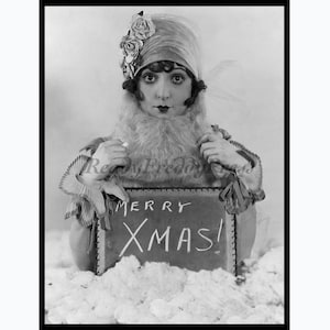 May include: Black and white vintage photograph of a woman wearing a cloche hat adorned with flowers and a fur collar. She holds a sign that reads "MERRY XMAS!" in chalk-style lettering, surrounded by a snowy setting.