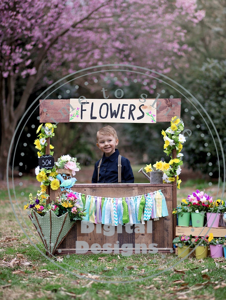 May include: A child stands behind a rustic wooden flower stall with a sign that reads "FLOWERS". The stall is decorated with colorful flowers, a price tag of 50p, and a banner. The background features a blurred pink flowering tree.