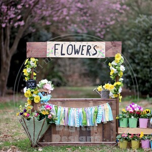 May include: A wooden flower stall with a sign saying "FLOWERS". The stall is adorned with various colorful flowers, a banner, and an umbrella. A small chalkboard displays the price of 50p. The background shows a blurred pink blossom tree.