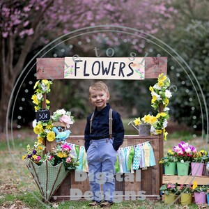 May include: A young boy poses in front of a rustic wooden flower stall with the word "FLOWERS" painted on it. The stall is decorated with colourful flowers, a chalkboard sign, and a green umbrella. The boy wears a dark blue shirt, light blue trousers, and braces.