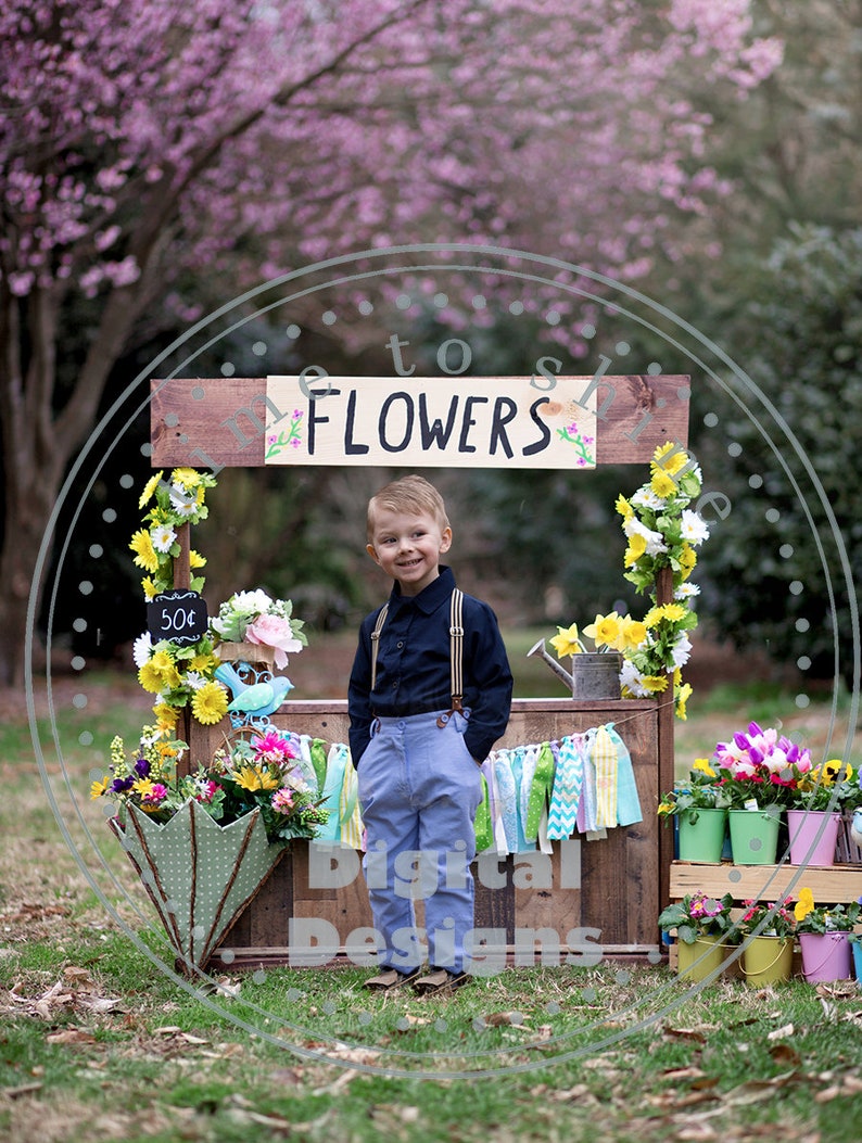 May include: A young boy poses in front of a rustic wooden flower stall with the word "FLOWERS" painted on it. The stall is decorated with colourful flowers, a chalkboard sign, and a green umbrella. The boy wears a dark blue shirt, light blue trousers, and braces.
