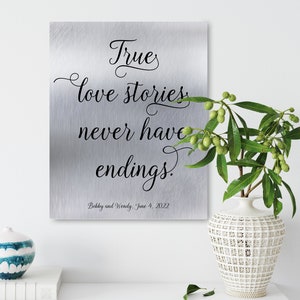 May include: A silver metal sign with a quote about love stories. The quote reads "True love stories never have endings." The sign also includes the names "Bobby and Wendy" and the date "June 4, 2022".