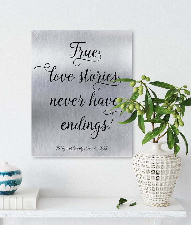 May include: A silver metal sign with a quote about love stories. The quote reads "True love stories never have endings." The sign also includes the names "Bobby and Wendy" and the date "June 4, 2022".