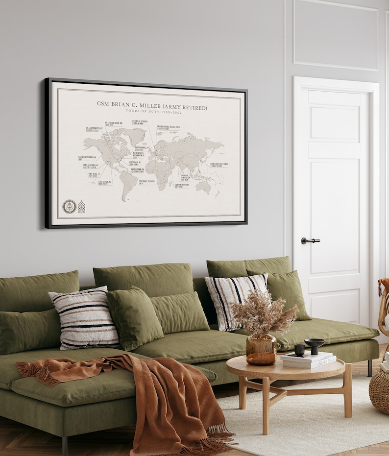 May include: A framed world map with pins marking locations around the globe. The map is titled "CSM Brian C. Miller (Army Retired) Tours of Duty 1993-2022". The map is hanging on a wall in a living room with a green sectional sofa and a wooden coffee table.