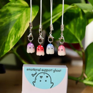 Op de afbeelding: Vijf kleurrijke spookvormige bedels met witte koorden en zilveren sluitingen. De bedels zijn in tinten paars, rood, blauw en roze. De bedels zijn gelabeld "emotionele steun spook" en "Boo!" met een cartoon spook op een blauwe achtergrond.