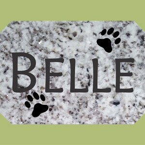 May include: A white granite pet memorial with the name "BELLE" engraved in black lettering. Three black paw prints are also engraved on the stone.