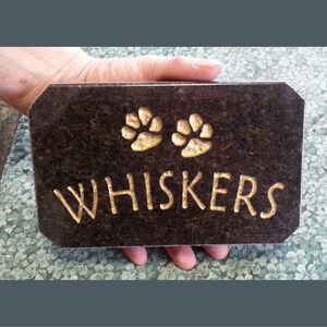 May include: A black granite pet memorial plaque with gold paw prints and the name "WHISKERS" engraved in gold.