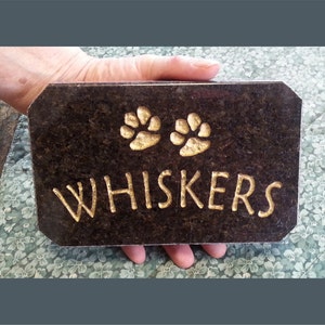 May include: A black granite pet memorial plaque with gold lettering that reads "WHISKERS" and two gold paw prints.