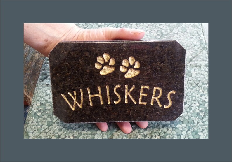 May include: A black granite pet memorial plaque with gold lettering that reads "WHISKERS" and two gold paw prints.