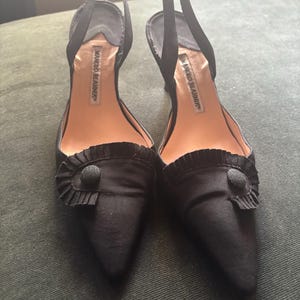 Puede incluir: Un par de zapatos de tacón negros con correa trasera y punta afilada. Los zapatos presentan un detalle de volantes en la parte delantera y un botón. La marca "MANOLO BLAHNIK" es visible en la plantilla.