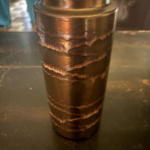 May include: A tall, cylindrical vase with a textured, layered design. The vase has a dark brown, metallic finish with lighter brown accents. The top of the vase has a slightly wider rim.