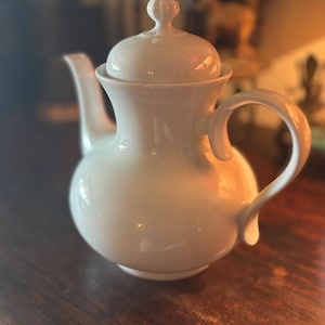 May include: A white ceramic teapot with a rounded body, a curved spout, and a loop handle. The lid has a small, decorative knob. The teapot is sitting on a dark wooden surface.