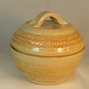 May include: A ceramic butter dish with a lid and a handle. The dish is a light beige color with a brown glaze.