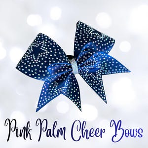 May include: A navy blue cheer bow with sparkling rhinestones arranged in star patterns. The bow features a central rhinestone band and is labeled "Pink Palm Cheer Bows" in cursive script.