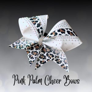 Könnte beinhalten: Eine große weiße Cheerleading-Schleife mit Leopardenmuster und Strassakzenten. Die Schleife hat eine funkelnde Strassmitte und ist mit "Pink Palm Cheer Bows" beschriftet.