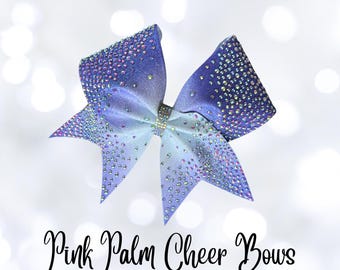 May include: A large, ombre blue cheer bow with iridescent rhinestones. The bow has a gradient effect, transitioning from light blue to a darker shade. The text "Pink Palm Cheer Bows" is written in cursive font.