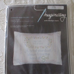 May include: A packaged cross-stitch kit for a baby pillow. The pillow design features the text "A bit of stardust blown from the hand of God" in blue and green, surrounded by a floral border. The pillow measures 10" x 14".