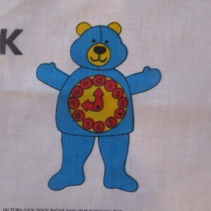May include: A blue bear-shaped clock design on a white fabric. The bear has a clock face on its belly with the hands pointing to one o'clock. The bear has yellow ears and a yellow nose. The text "TICK-TOCK" is visible.