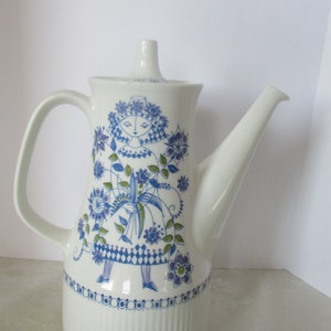 May include: White ceramic coffee pot with a blue and green floral design featuring a stylized woman in a blue dress. The pot has a ribbed base and a spout on the right side.