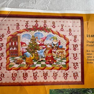 May include: A framed cross-stitch kit featuring a Christmas scene with elves decorating a tree. The design includes a house interior with elves, a Christmas tree, and the numbers 1-24. Finished size is 18 inches.