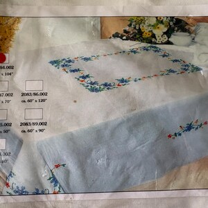 Op de afbeelding: Een verpakte tafelkleed met een geborduurd bloemenontwerp. Het tafelkleed is wit met een lichtblauwe rand en heeft blauwe en rode bloemborduurwerk. De verpakking bevat afmetingen van ongeveer 152 cm x 264 cm, 127 cm x 178 cm, 152 cm x 305 cm, 127 cm x 127 cm, 152 cm x 229 cm en 152 cm x 152 cm.