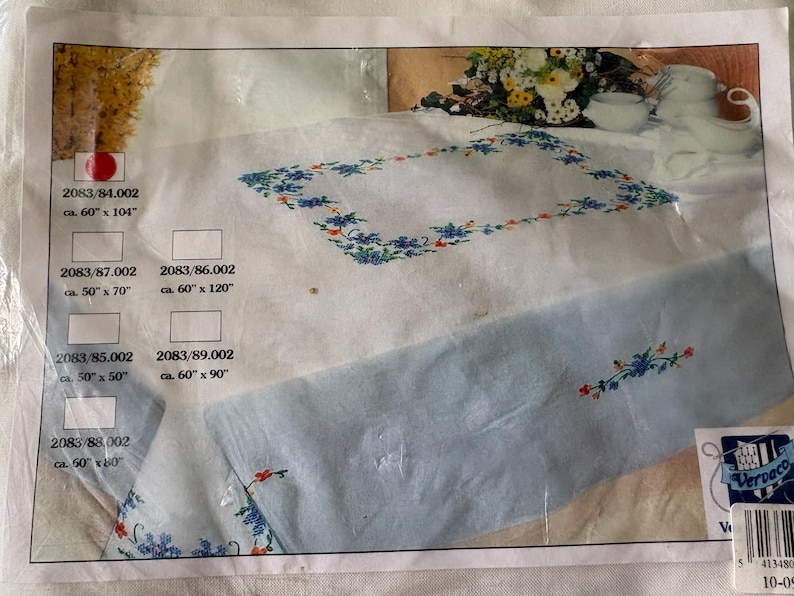 Op de afbeelding: Een verpakte tafelkleed met een geborduurd bloemenontwerp. Het tafelkleed is wit met een lichtblauwe rand en heeft blauwe en rode bloemborduurwerk. De verpakking bevat afmetingen van ongeveer 152 cm x 264 cm, 127 cm x 178 cm, 152 cm x 305 cm, 127 cm x 127 cm, 152 cm x 229 cm en 152 cm x 152 cm.