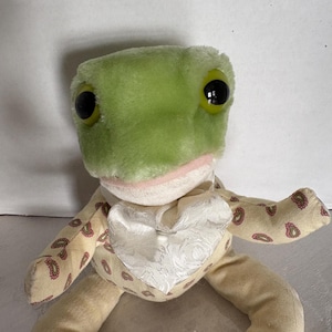 Peut inclure: Une peluche en forme de grenouille avec une tête vert clair, de grands yeux noirs et une bouche rose. Elle a des bras et des jambes crème avec un motif cachemire. La grenouille porte un gilet blanc. L'étiquette indique "eden".