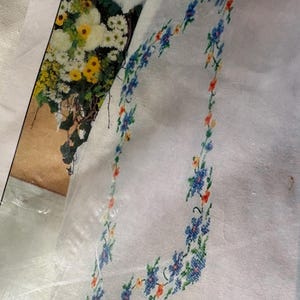 Op de afbeelding: Witte stof met een bloemenkruissteekrand in blauw, oranje en geel. Het ontwerp heeft kleine bloemen en groene bladeren. Op de achtergrond is een afbeelding van een boeket bloemen te zien.
