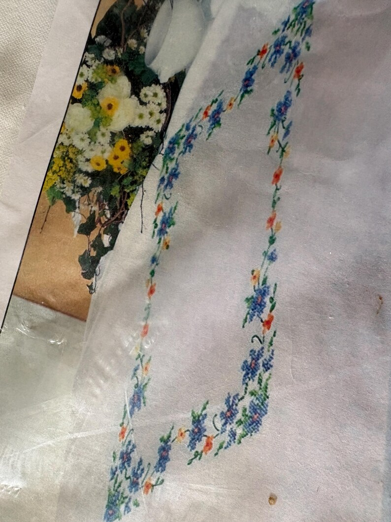 Op de afbeelding: Witte stof met een bloemenkruissteekrand in blauw, oranje en geel. Het ontwerp heeft kleine bloemen en groene bladeren. Op de achtergrond is een afbeelding van een boeket bloemen te zien.