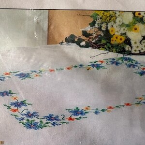 Op de afbeelding: Wit tafelkleed met een geborduurde bloemenrand in blauw, oranje en groen. Op de achtergrond staat een boeket witte en gele bloemen. De geborduurde rand vormt een rechthoek.